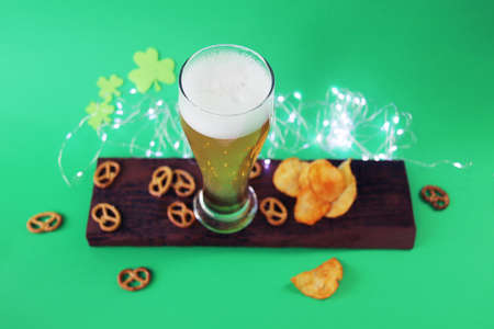 Patrick day, fresh foamy beer in a glass, chips and crackers on a wooden board, shamrock on a green...