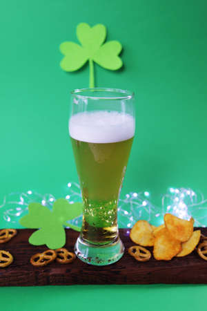 Patrick day, fresh foamy beer in a glass, chips and crackers on a wooden board, shamrock on a green background, party, congratulations, postcardの写真素材