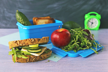 Fresh vegetables, fruits, cheese and micro greens for preparing a school lunch and packaging in a box, on a table, against the background of an alarm clock and a chalkboardの写真素材