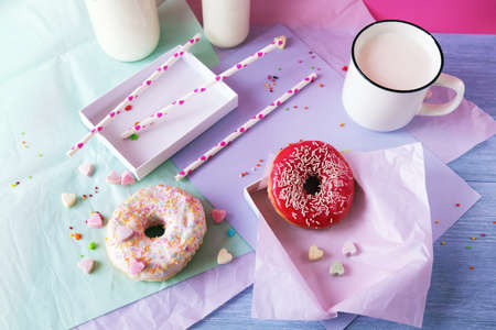 Freshly baked donuts glazed with various pink shades and sprinkled with caramel hearts, milk and yogurt in a cup and glass bottles, lilac-colored paper on the tableの写真素材