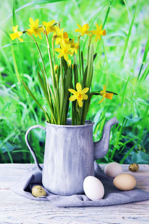 Easter eggs, metal vintage jug with a bouquet of fresh blooming yellow daffodils on a wooden table against the background of green grass, congratulations concept, village, farmの写真素材
