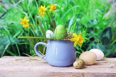 Easter eggs, a cup with Easter decorations and a flower, on a wooden table, against the background of green grass and blooming yellow daffodils, the concept of home celebrations, congratulations, postの写真素材