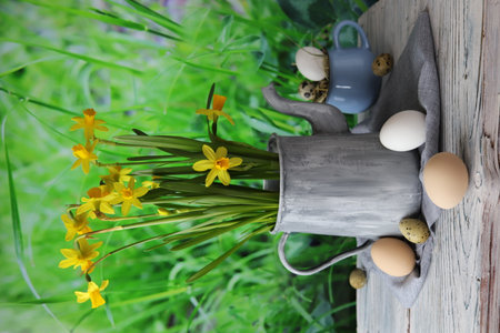 Easter eggs, metal vintage jug with a bouquet of fresh blooming yellow daffodils on a wooden table against the background of green grass, congratulations concept, village, farmの写真素材
