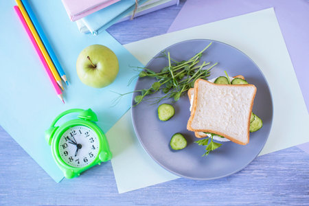 School lunch with sandwiches, alarm clock and stationery on color wooden background