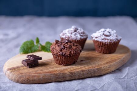 Chocolate muffins on a wooden board, strewed with icing sugar. Homemade baking. In the background is a cup of coffee and a plate with muffins. Wooden background. Selective focus, close up.の写真素材