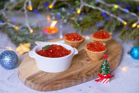 Red caviar (salmon caviar) in a white bowl on a wooden board. In the background are tartlets with caviar, Christmas tree branches, toys, candle and bokeh.の写真素材