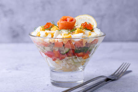 Layered salad with salted trout (salmon), caviar, eggs and vegetables. Traditional dish, one portion in a glass goblet on a gray background. Close-up.の写真素材