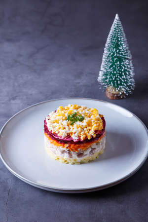 Herring salad under a fur coat. Traditional Russian multilayered salad from herring, beets, potatoes, carrots and eggs. Close-up, gray background, miniature Christmas tree.の写真素材