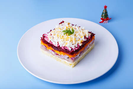 Herring salad under a fur coat. Traditional Russian multilayered salad from herring, beets, potatoes, carrots and eggs. Close-up, blue background, miniature Christmas tree.の写真素材