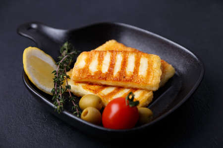 Grilled halloumi cheese in a black pan with olives, tomatoes, cucumbers, thyme and lemon. Traditional cheese of the Eastern Mediterranean. Black background, selective focus, close-up.の写真素材