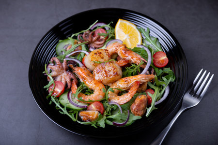 Salad with seafood, arugula, tomatoes, cucumbers, red onion and lemon on a black plate. Black background, close-up.の写真素材