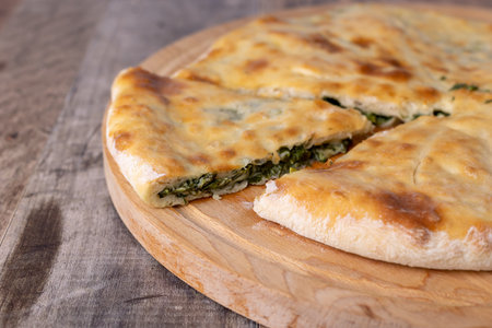 Pie with spinach, herbs and suluguni cheese on a wooden board. Whole pie with cut off piece. Traditional round flat pie with filling. Homemade pastries of Caucasian and Balkarian cuisine. Selective focus, close-up.の写真素材