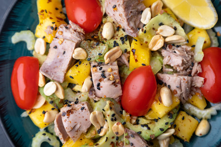 Salad with canned tuna, tomatoes, peanuts, mango, avocado, celery, chili flakes and black sesame. Light summer salad with olive oil and a lemon lobule. Home cooking. Selective focus, close-up, black background.の写真素材
