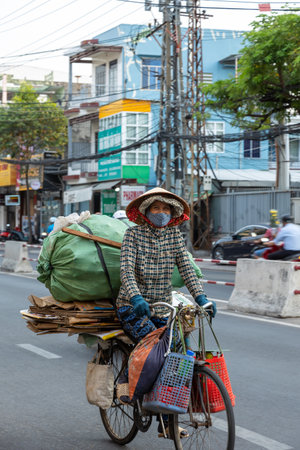 Nha Trang, Vietnam - April 22, 2024: A Vietnamese elderly woman in traditional closed clothes and hat carries trash on an old bicycle along the city street and smiles. Selective focus.のeditorial素材