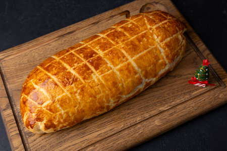 Whole beef Wellington on a wooden board. A small toy Christmas tree nearby. Traditional festive dish of beef tenderloin with mushrooms and Parma ham baked in puff pastry. Selective focus, close-up.の写真素材