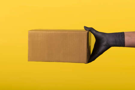Online shopping and express delivery. Courier hand in medical rubber gloves holding brown box isolated on colored background with copy space. Fast and free delivery by vehicle. Quarantine Close-up.の写真素材