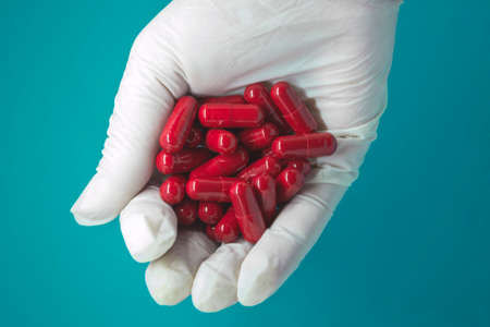 A hand in a medical glove holds a handful of red pills. On a blue background. Pharmaceutical...