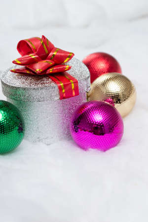 On the snow there is a silver gift box with a red bow and glass multi-colored Christmas decorations. New year concept, new year card.Vertical photoの写真素材