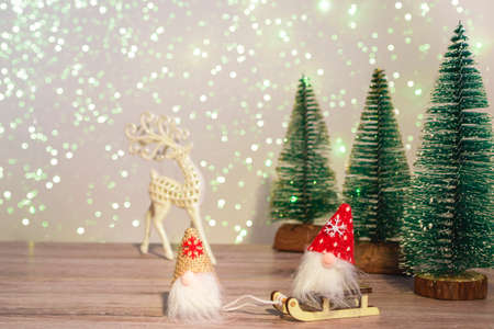 A gnome in a white hat leads a gnome in a red hat on a sleigh. Winter background with Christmas trees in sparkles and a deer. Happy Holidays.の写真素材