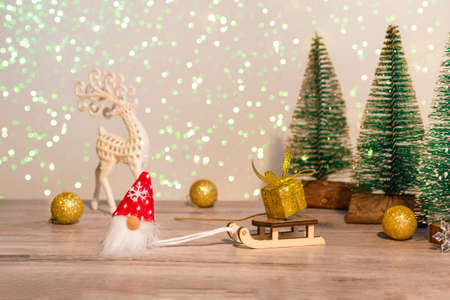 A gnome in a red hat brought a golden gift on a sleigh. Winter background Christmas trees in sparkles and a deer. Happy Holidays.の写真素材