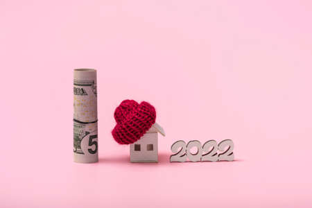 The concept of the heating season 2022 or purchase. On a pink background, a wooden house in a red hat and a stack of dollar bills close up with copy spaceの写真素材