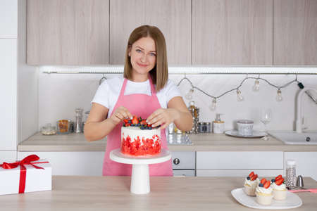 Pastry chef confectioner young caucasian woman in the kitchen. She is decorating the cake on a stand. Nearby are cakesの写真素材