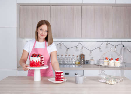 Pastry chef confectioner young caucasian woman in the kitchen. In front of her is a cake on a stand. She holds out a piece of cake on a plate. Near sweet cupcakes, a cup and sugar in a sugar bowlの写真素材