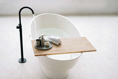 White bathtub fills with foam water in a modern apartment with stylish loft-style interior design,...