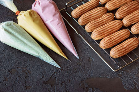 French pastry dessert eclair. Freshly baked aromatic custard cakes cool on the wire rack. Pastry bags with mint, mango, berry creams. Soft selective focus.の写真素材