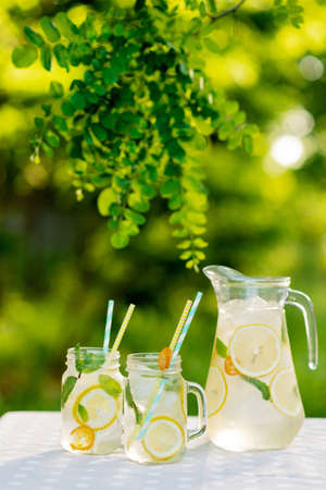 Lemonade refreshing drink in a jug and jars with lemons, fresh mint and ice on a garden table. Summer picnic outdoors. Soft selective focus.の写真素材