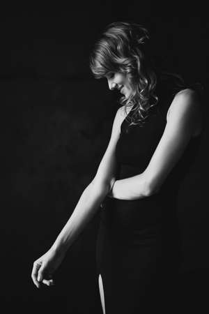 Beautiful elegant slim woman in a black dress in a loft studio modestly looks down. Soft selective focus. Black and white art photo.の写真素材