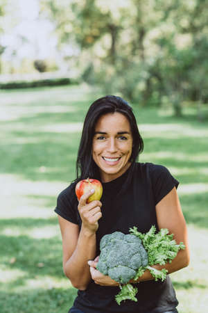 Portrait of smiling young tanned woman in the park holding an apple and a bouquet of greens with...