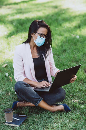 Young woman with protective mask using laptop in a public park while working outdoors. Distance learning and teleworking during a pandemic. Soft selective focus.の写真素材