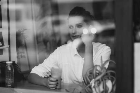A beautiful young girl sits in a cafe behind a glass with a cup of coffee and looks thoughtfully out the window. Black and white art photo through glass with reflection. Soft selective focus.の写真素材