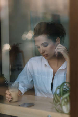 Beautiful young girl sits in a cafe behind a glass with a cup of coffee and and looks into a...