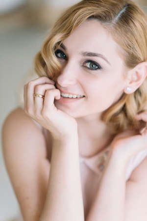 A beautiful blonde woman in an elegant powdery pink dress in the morning in a bright room playfully smiles close-up portrait. Soft selective focus.の写真素材