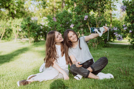 Smiling pretty young women influencer bloggers are filming or recording video with their smartphone on a stabilizer, in a sunny green park outside. Blogging concept. Soft selective focus.の写真素材