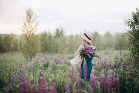 Beautiful romantic girl blonde in a dress and hat in a field with a bouquet of purple flowers of lupins at sunrise. Soft selective focus. Copy space.の写真素材