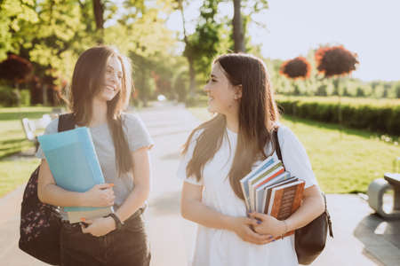 Two happy smiling student girls are walking and talking to each other on campus at sunset with warm light. Soft selective focus.の写真素材