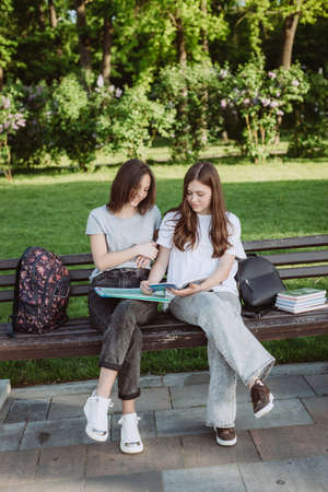 Two student girls are looking at an open book on a bench in the park. Distance education, preparation for exams. Soft selective focus.の写真素材