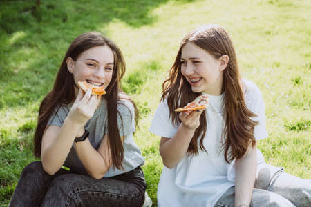 Two cheerful young teen friends in the park eating pizza. Women eat fast food. Not a healthy diet....