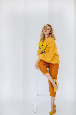 Happy young blonde woman with a bouquet of yellow roses in a voluminous sweater posing on a white background in the studio. Soft selective focus.の写真素材