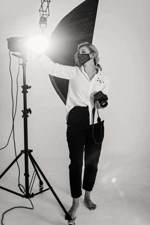 A beautiful woman photographer in a protective mask works in a professional photo studio among the lighting equipment. Hobby profession. Soft selective focus.の写真素材