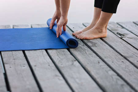 woman twists yoga mat outdoors near the river on a wooden pier in the morning. Healthy lifestyle concept. Morning meditation, relaxation.の写真素材
