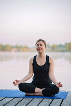 A 36-year-old young Caucasian woman practices yoga in the lotus position outdoors near the river on a wooden pier in the morning. Healthy lifestyle concept. Morning meditation, relaxation.の写真素材