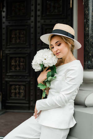 Young stylish lady in a white trouser suit and a hat with an elegant hairstyle with a bouquet of hydrangea flowers on the streets of the city. The woman is the bride.の写真素材