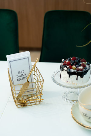 An exquisite berry cake on a glass stand on a table with a vase of flowers and a tea set in a modern...