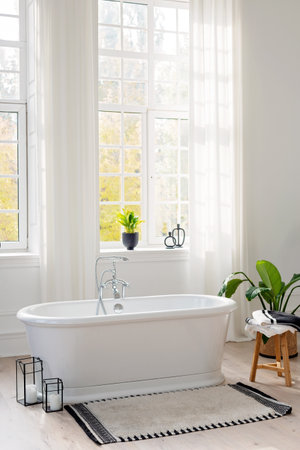 Stylish modern bathroom interior. Vertical view of an empty free-standing bathtub on a wooden floor in a bright room against the backdrop of a large window and green houseplants. Soft selective focus.の写真素材