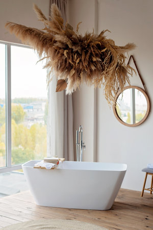 Comfortable bright bathroom with a boho-chic interior design, a free-standing white bath against the background of a panoramic window. hanging decor from dry grass. Soft selective focus.の写真素材