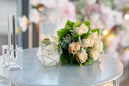 A pair of beautiful wedding rings in a modern glass stand and a delicate bridal bouquet of roses. Marriage, ceremony. Soft selective focus.の写真素材